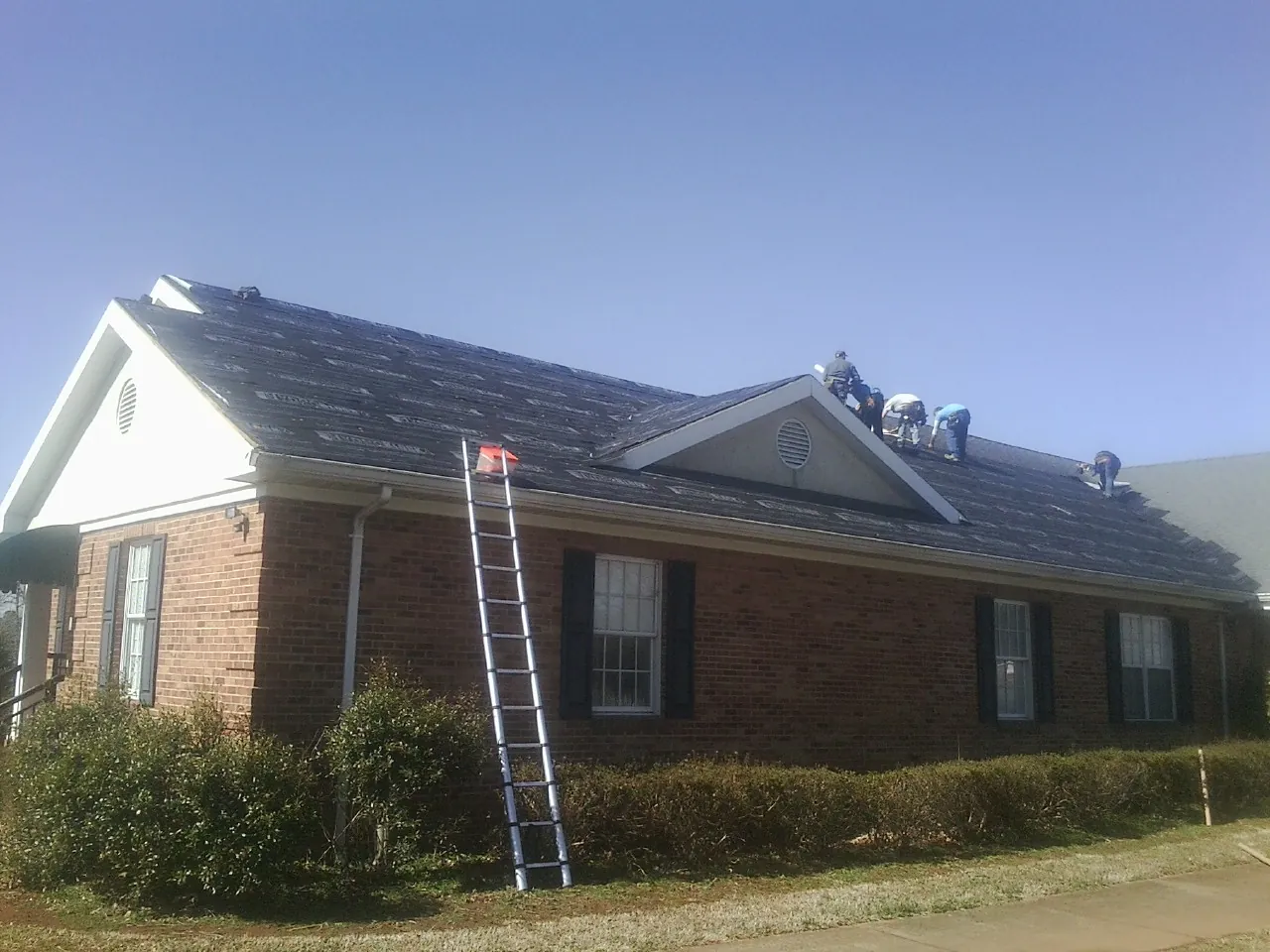 Crew tearing shingles off a brick ranch roof with a ladder set — Metal Roof Repair work by our crew in Old Monroe