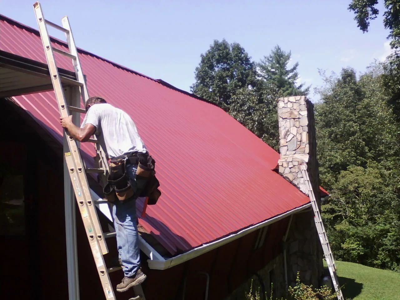 Roofer on a ladder beside a red metal roof and stone chimney — Soffit & Fascia Repair by Roofing Brothers in Old Monroe
